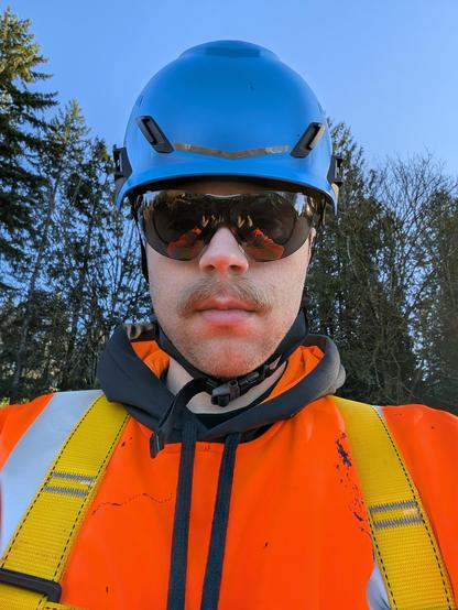 Selfie of me, a white male with a light colored moustache. I'm standing in front of tall pine trees silhouetted against a blue sky, implying I'm standing on a roof or somewhere high up. I'm wearing sunglasses, an orange hi-vis sweater, a blue MSA helmet, and a yellow fall protection harness
