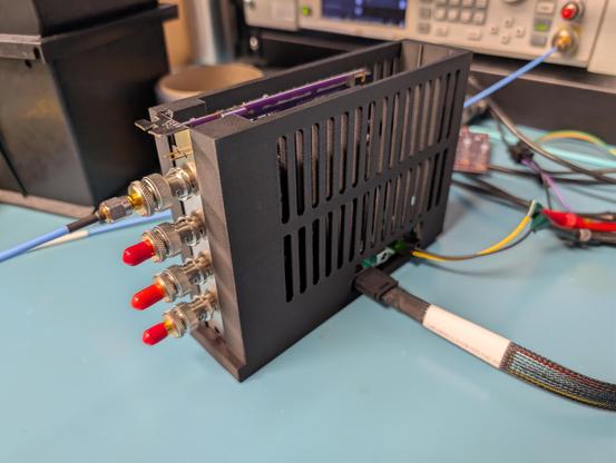 PCIe card on a blue ESD mat in a black vented enclosure with a 12V power connection and PCIe data connection coming off the bottom right side and four BNCs on the back